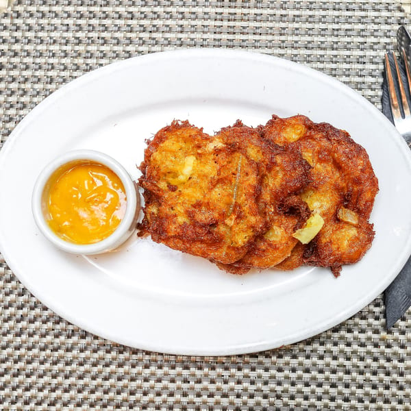 Three golden-brown Jamaican fritters with a side of orange dipping sauce on a white plate.