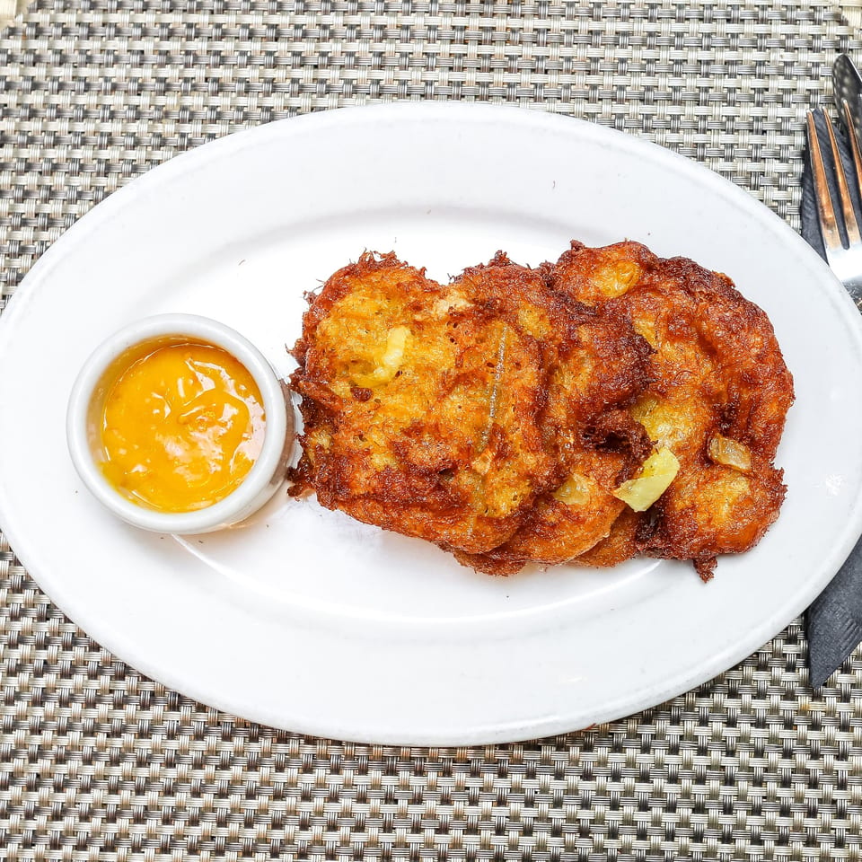Three golden-brown Jamaican fritters with a side of orange dipping sauce on a white plate.