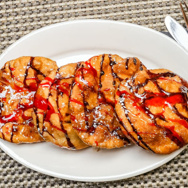 Four golden-brown Jamaican fried dumplings, drizzled with red and chocolate syrup, dusted with powdered sugar.