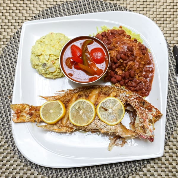 Whole fried fish with lemon slices, potato salad, rice and beans, and a side of stewed vegetables.