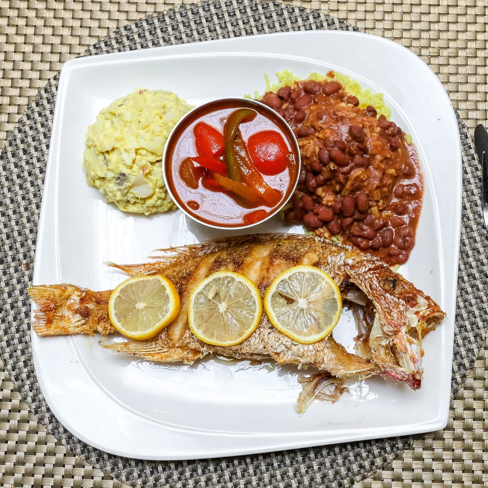 Whole fried fish with lemon slices, potato salad, rice and beans, and a side of stewed vegetables.